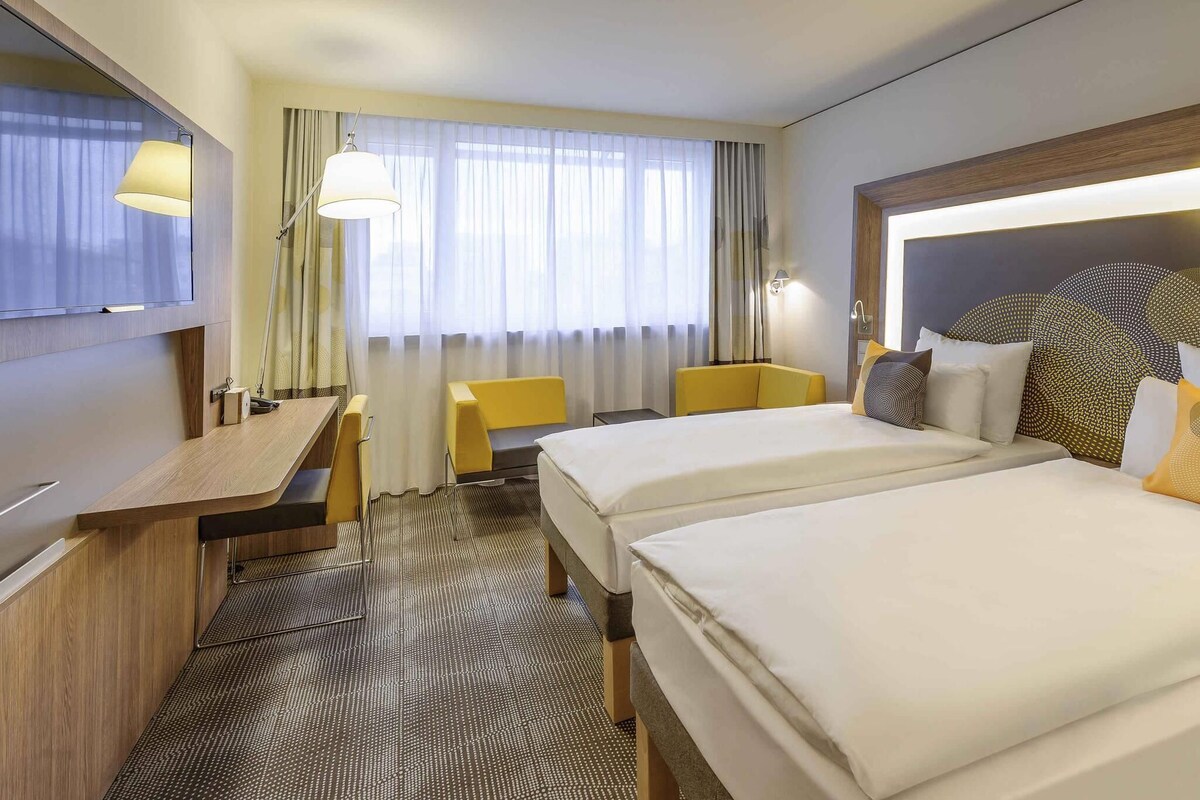 A hotel room with two beds, a desk, a chair, a TV, and a window with curtains.