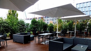 An outdoor patio area with wooden flooring, black and grey seating, and large white umbrellas providing shade. - Novotel München City Arnulfpark (Munich)