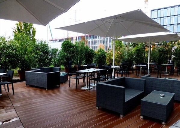 Un patio al aire libre con suelo de madera, asientos negros y grises y grandes sombrillas blancas que dan sombra.