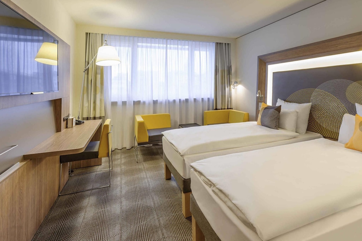 Een hotelkamer met twee bedden, een bureau, een stoel, een televisie en een raam met gordijnen.