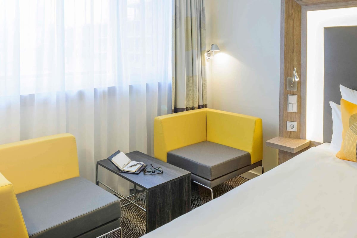 Hotelkamer met een bed, twee gele fauteuils, een klein tafeltje met een notitieboekje en glazen, een lamp en een lichtschakelaar aan de muur.