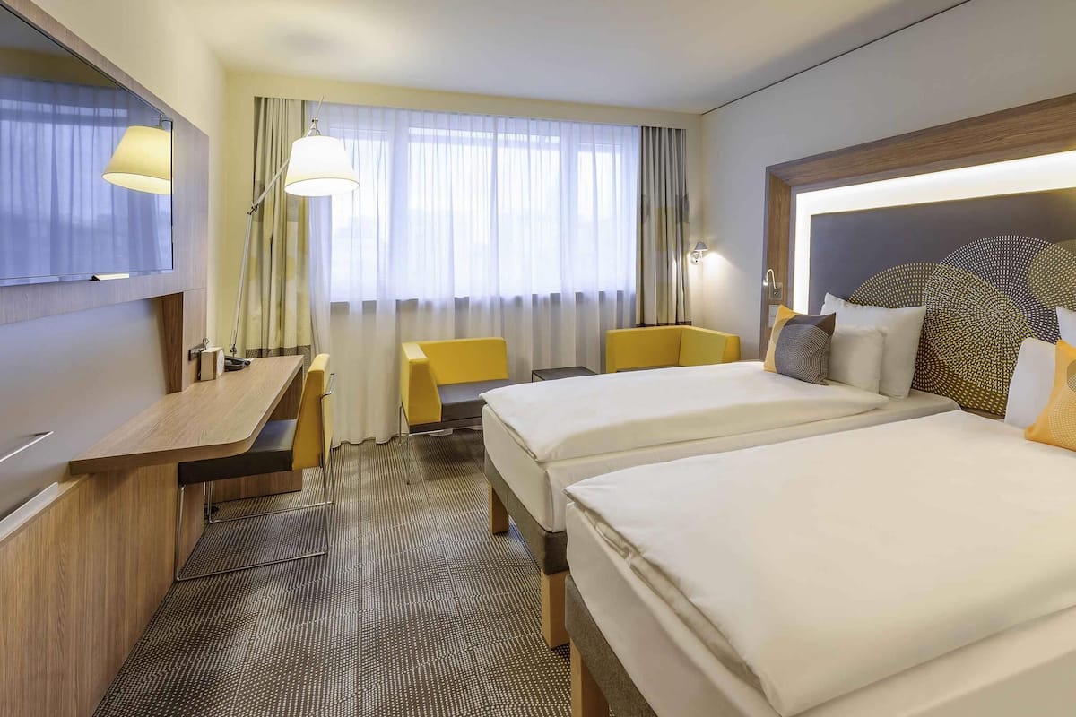 A hotel room with two beds, a desk, a chair, a TV, and a window with curtains.