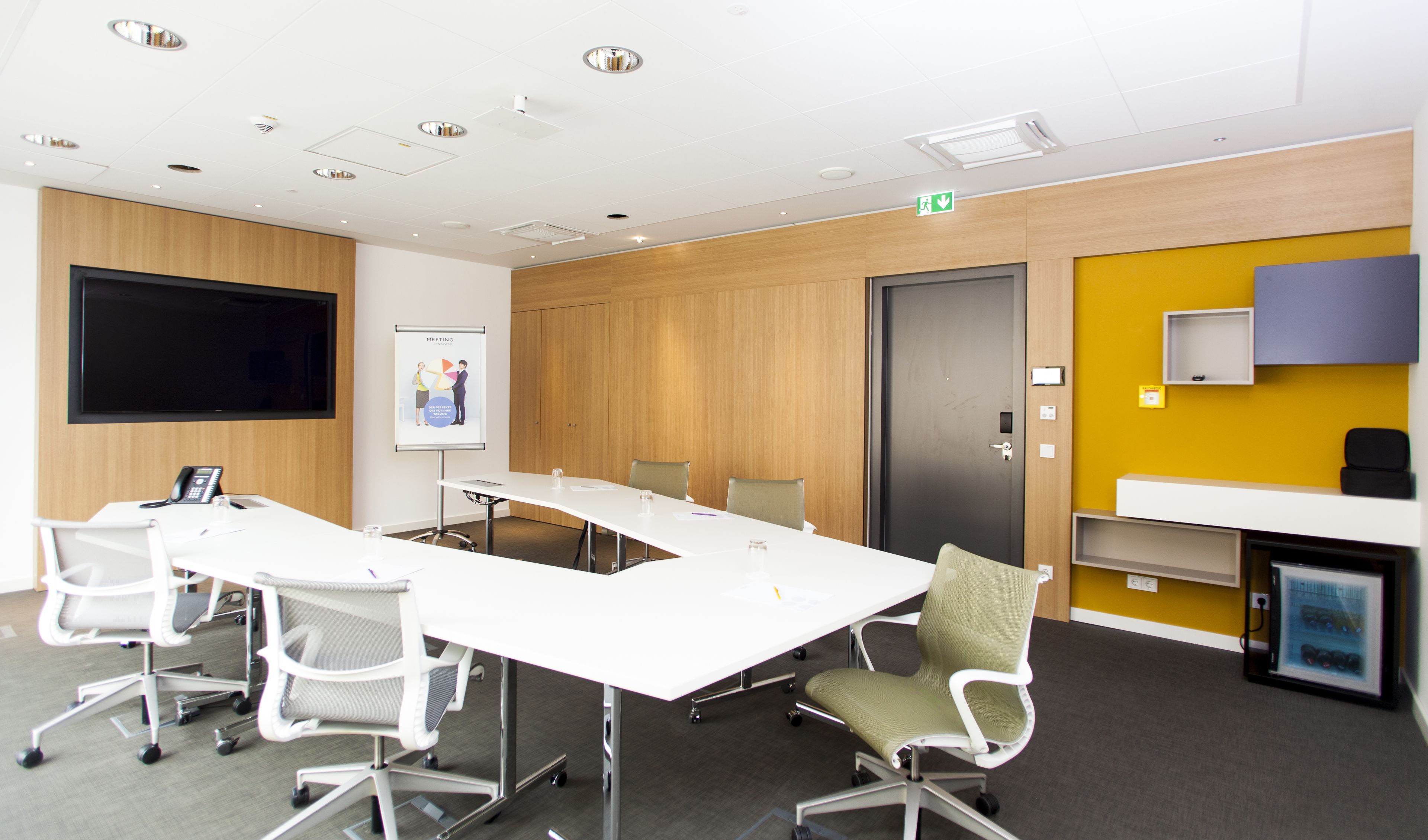 A modern meeting room with a long table, chairs, a projector screen, and a wooden panel wall.