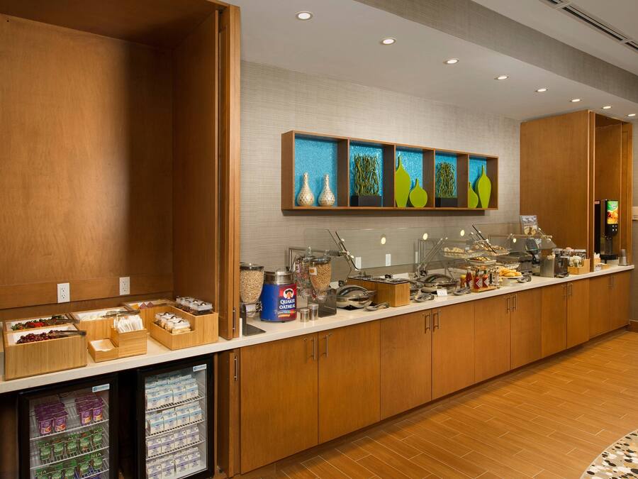 A hotel breakfast area with wooden cabinets, a refrigerated display case, and a variety of food items.