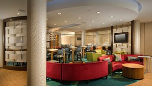 A modern hotel lobby with a curved ceiling, a seating area with red and green sofas, a dining area with tables and chairs, and a flat-screen TV mounted on the wall. - SpringHill Suites Bridgeport Clarksburg (Bridgeport)