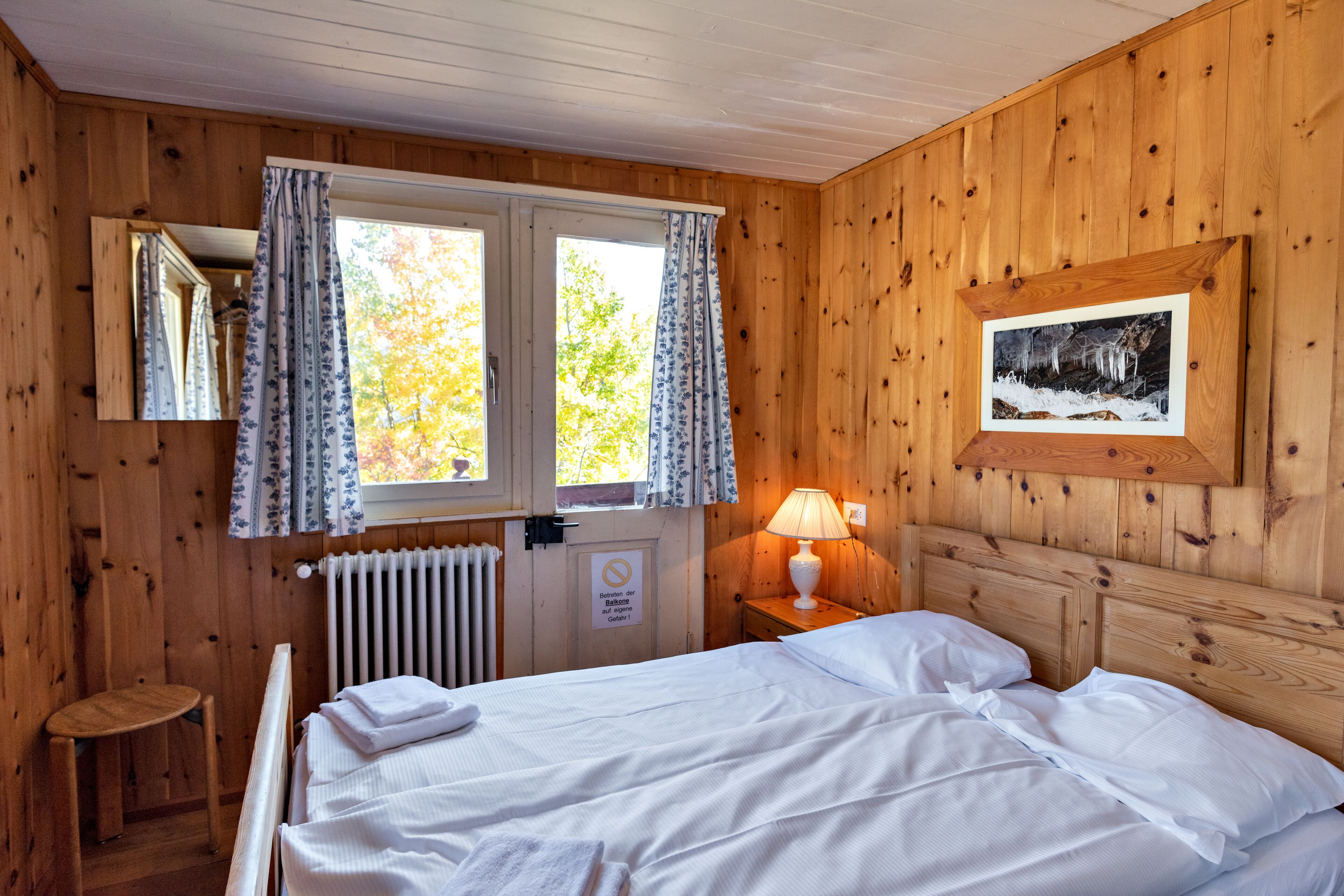 A wooden cabin room with a bed, a window with curtains, a radiator, a lamp, and a framed picture on the wall.
