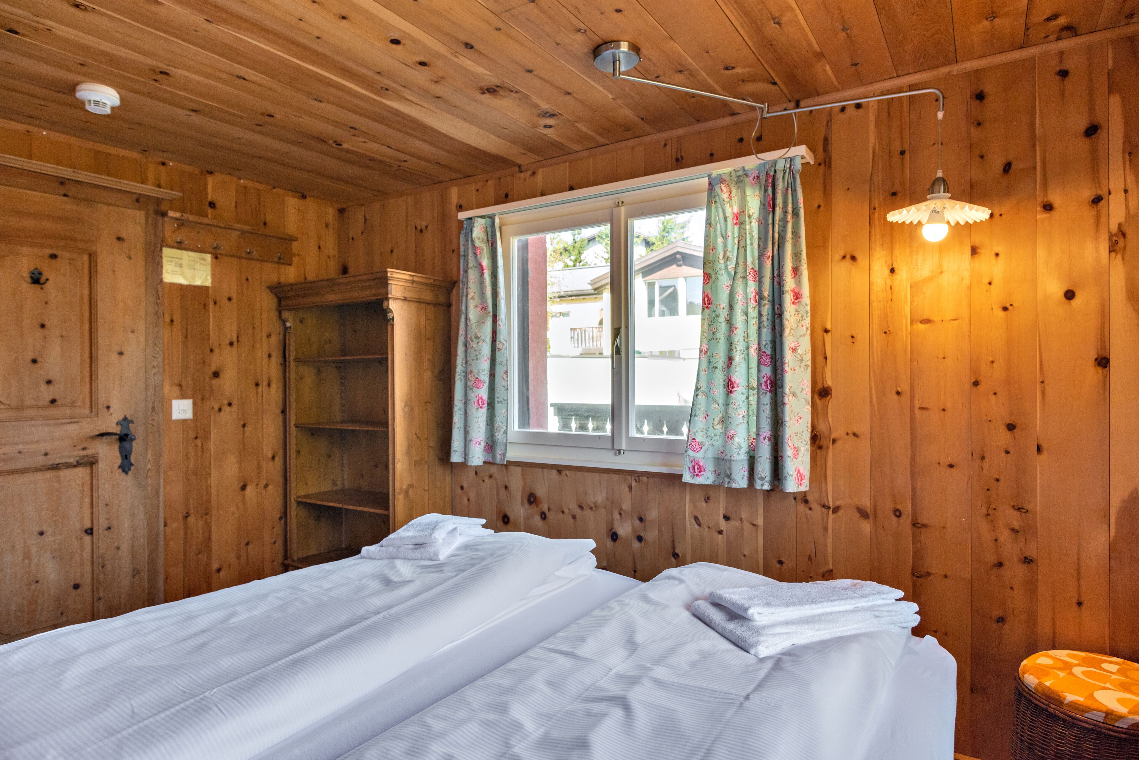 A wooden cabin room with two beds, a window with floral curtains, a ceiling light, and a wooden door.