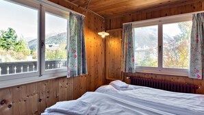 Een kamer met houten lambrisering, een bed, twee ramen met bloemen gordijnen en uitzicht op bergen.