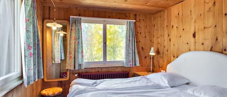 A wooden cabin room with a bed, a window with curtains, a radiator, and a small table with a lamp.