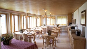 Een eetruimte met houten tafels en stoelen, grote ramen en hangende lampen.