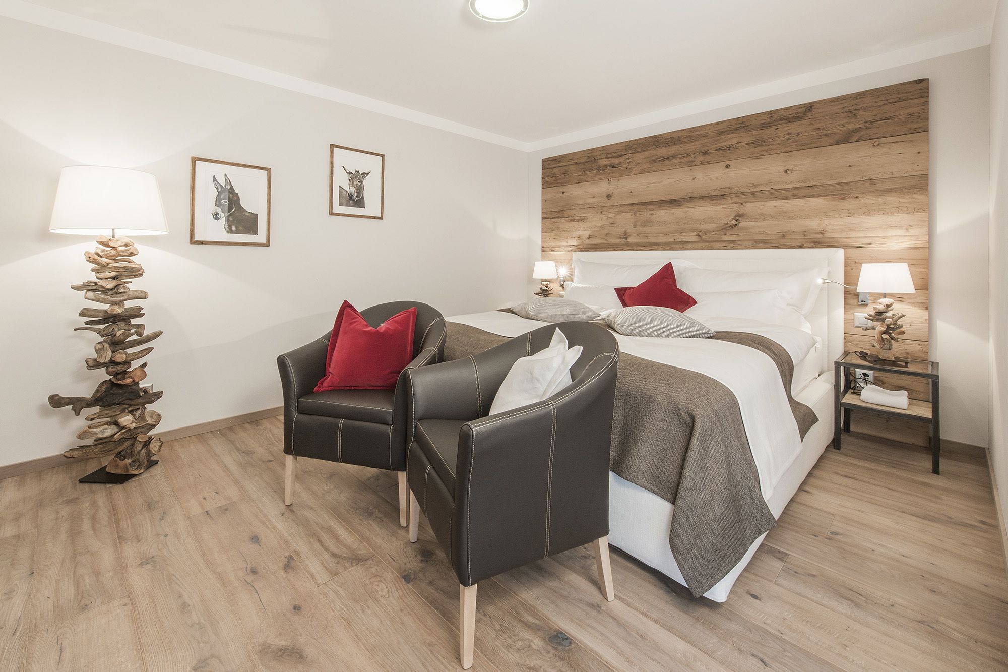 Una habitación de hotel moderna con cabecera de madera, cama con sábanas blancas y almohadas rojas, un sillón de cuero negro, suelo de madera y dos lámparas de noche.