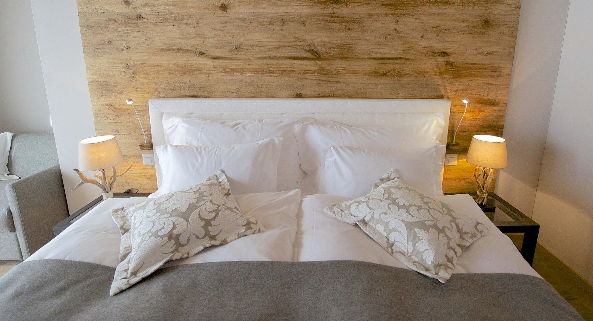 A neatly made bed with white linens and two patterned pillows, flanked by two bedside tables with lamps, against a wooden headboard.