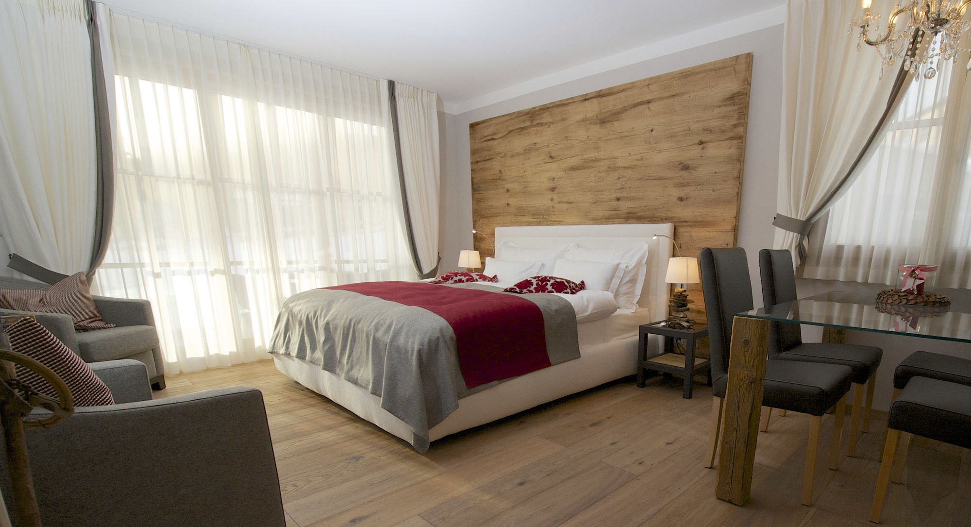 A hotel room with a wooden headboard, a bed with a red and grey bedspread, a grey sofa, a glass dining table with chairs, and a chandelier.