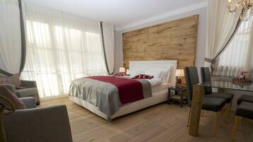 A hotel room with a wooden headboard, a bed with a red and grey bedspread, a grey sofa, a glass dining table with chairs, and a chandelier.