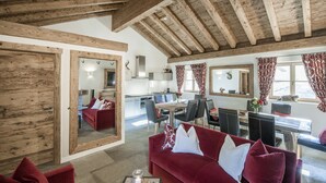 A cozy living area with a red sofa, wooden beams, a dining table, and a kitchen in the background. - Hotelino Petit Chalet (Celerina-Schlarigna)