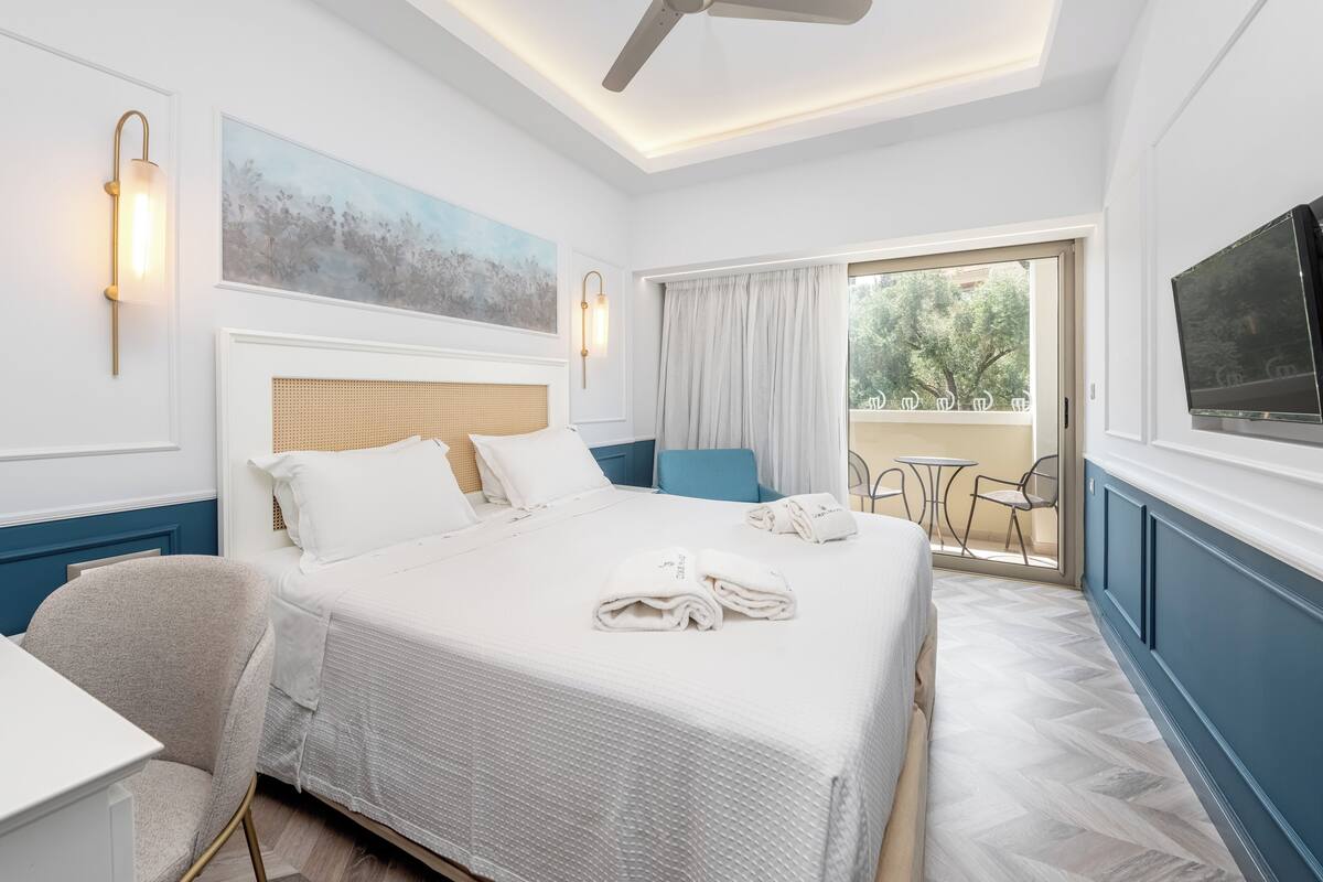 A hotel room with a bed, a desk, a chair, a ceiling fan, a painting, a window with curtains, and a TV.