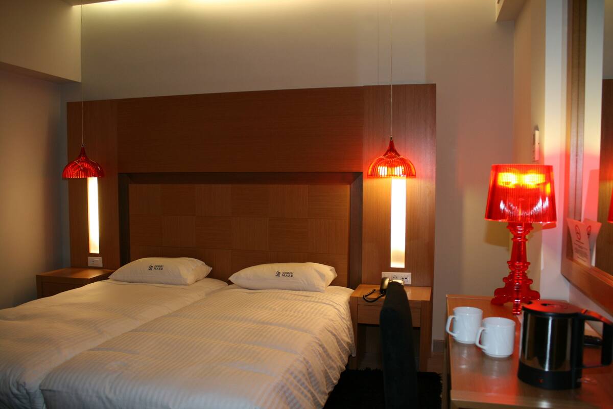 A hotel room with a wooden headboard, two beds, a nightstand with a red lamp, a coffee maker, and two white mugs.