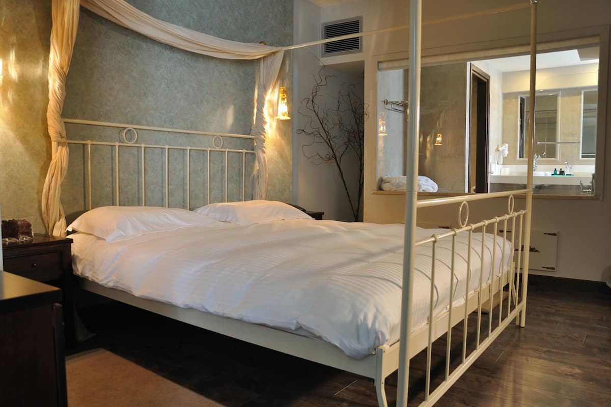 A neatly made bed in a room with a large window, a bedside table, and a mirror.