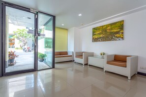 A modern lobby with a glass door entrance, two white sofas with brown cushions, a small table with a vase, and a large painting of a pond on the wall.