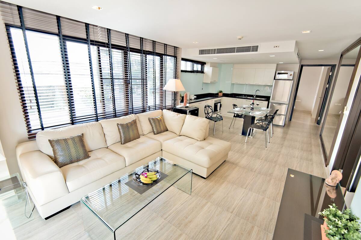 A modern living room with a beige sectional sofa, a glass coffee table with fruit, and a kitchen in the background.