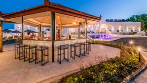 A covered outdoor bar area with seating, a pool, and a building with multiple balconies in the background. - Belmare Hotel (Rhodes)