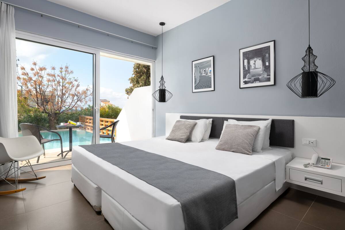 A modern hotel room with a large bed, a bedside table with a phone, and a view of a swimming pool and outdoor seating.