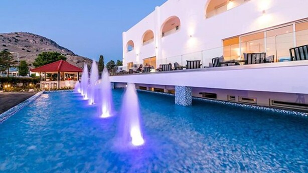 A modern hotel with a pool featuring water jets, a covered seating area, and a mountain backdrop. - Belmare Hotel (Rhodes)
