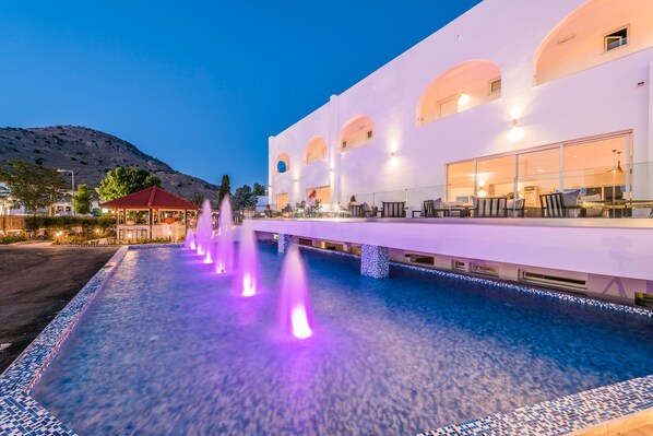A pool area with a fountain, a covered seating area, and a building with arched windows.