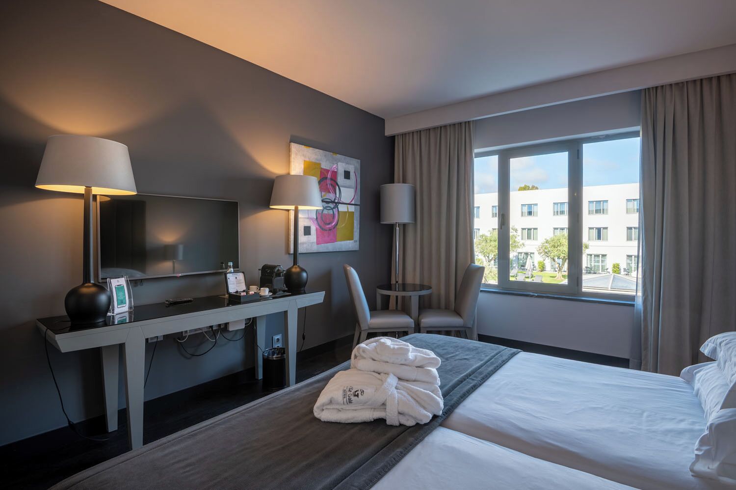 A hotel room with a bed, desk, chair, and a window with a view of buildings.
