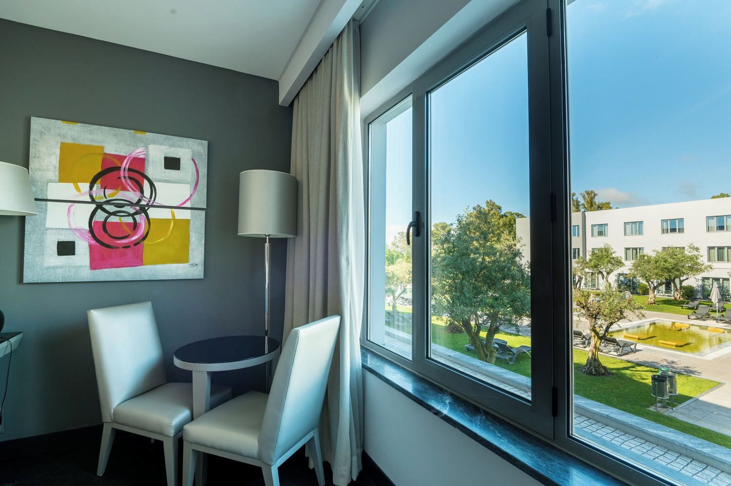 Un cuarto de hotel con un ventanal grande, una mesa redonda, dos sillas, una lámpara de pie y una obra de arte abstracto en la pared.