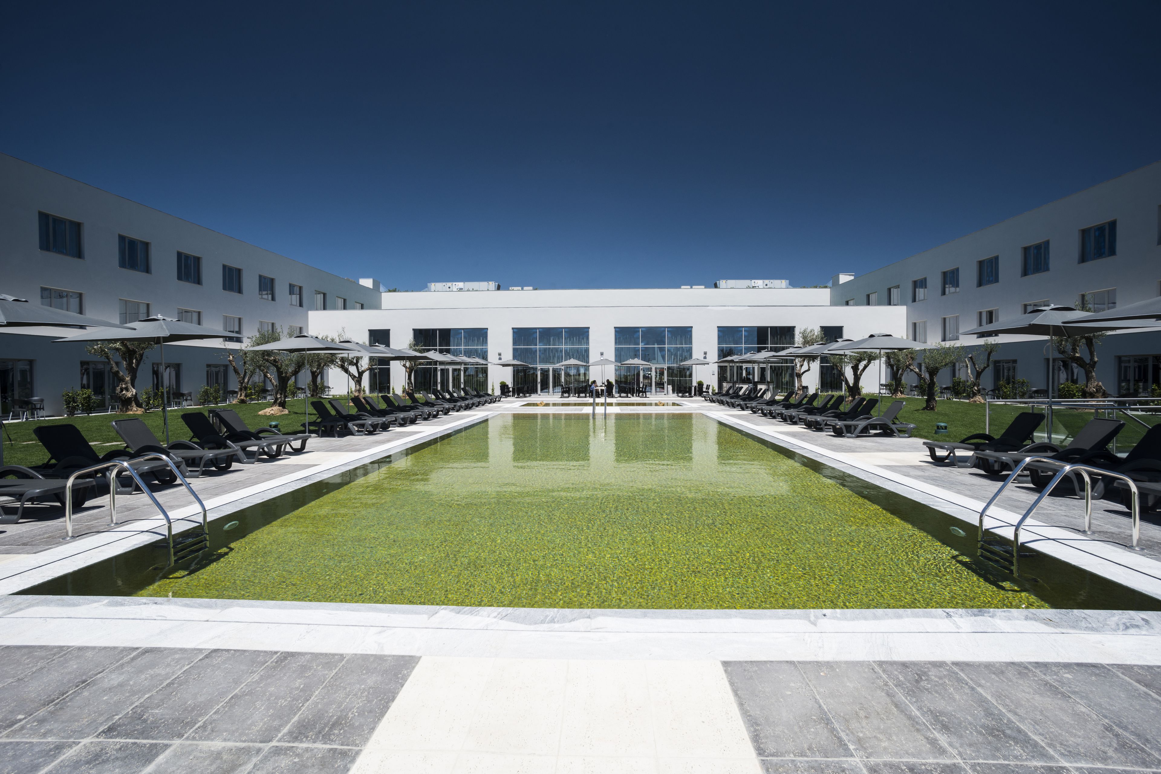 A modern hotel with a central swimming pool, sun loungers, and a clear blue sky.