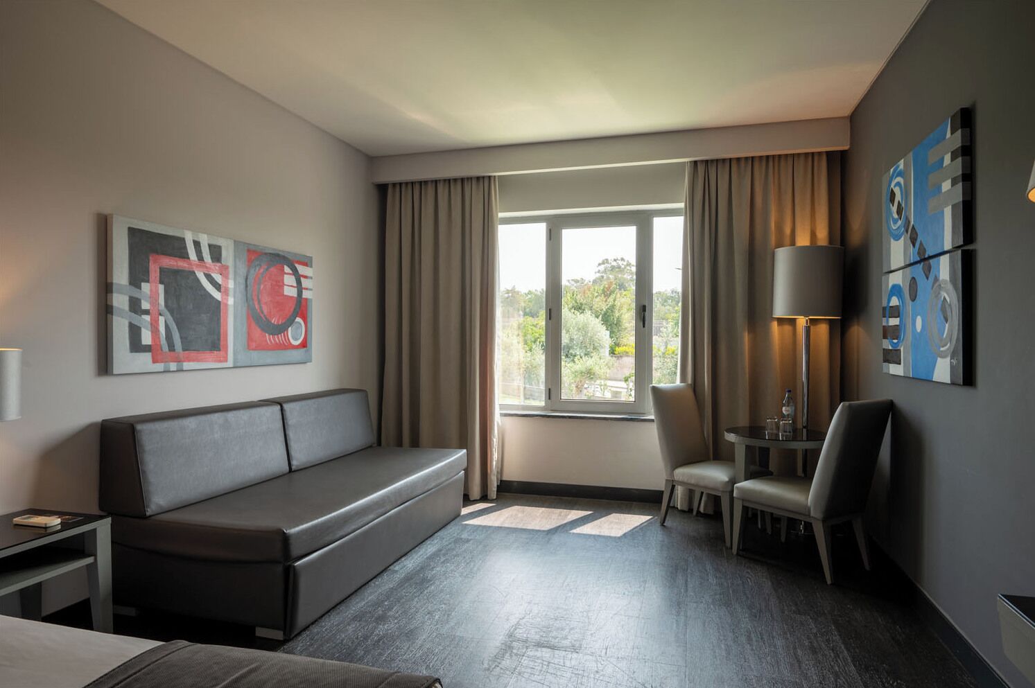 A hotel room with a sofa, a chair, a small table with a lamp, and abstract artwork on the walls.