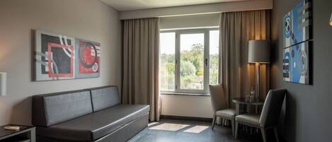A hotel room with a sofa, a chair, a small table with a lamp, and abstract artwork on the walls.