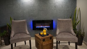A modern living room with two grey armchairs, a wooden coffee table with a tray of orange juice, and a flat-screen TV mounted on a textured grey wall.
