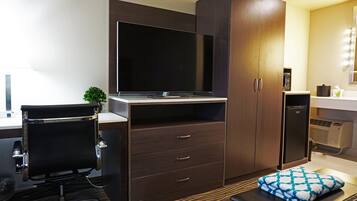 A modern hotel room with a flat-screen TV, a desk with a chair, a wardrobe, and a small kitchenette.
