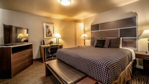 A hotel room with a large bed, a desk, a chair, a bench, a TV, and a wall-mounted headboard.