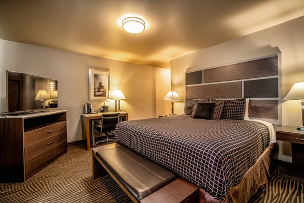 Habitación de hotel con una cama grande, un escritorio, una silla, un banco, un televisor y un cabecero fijado a la pared.