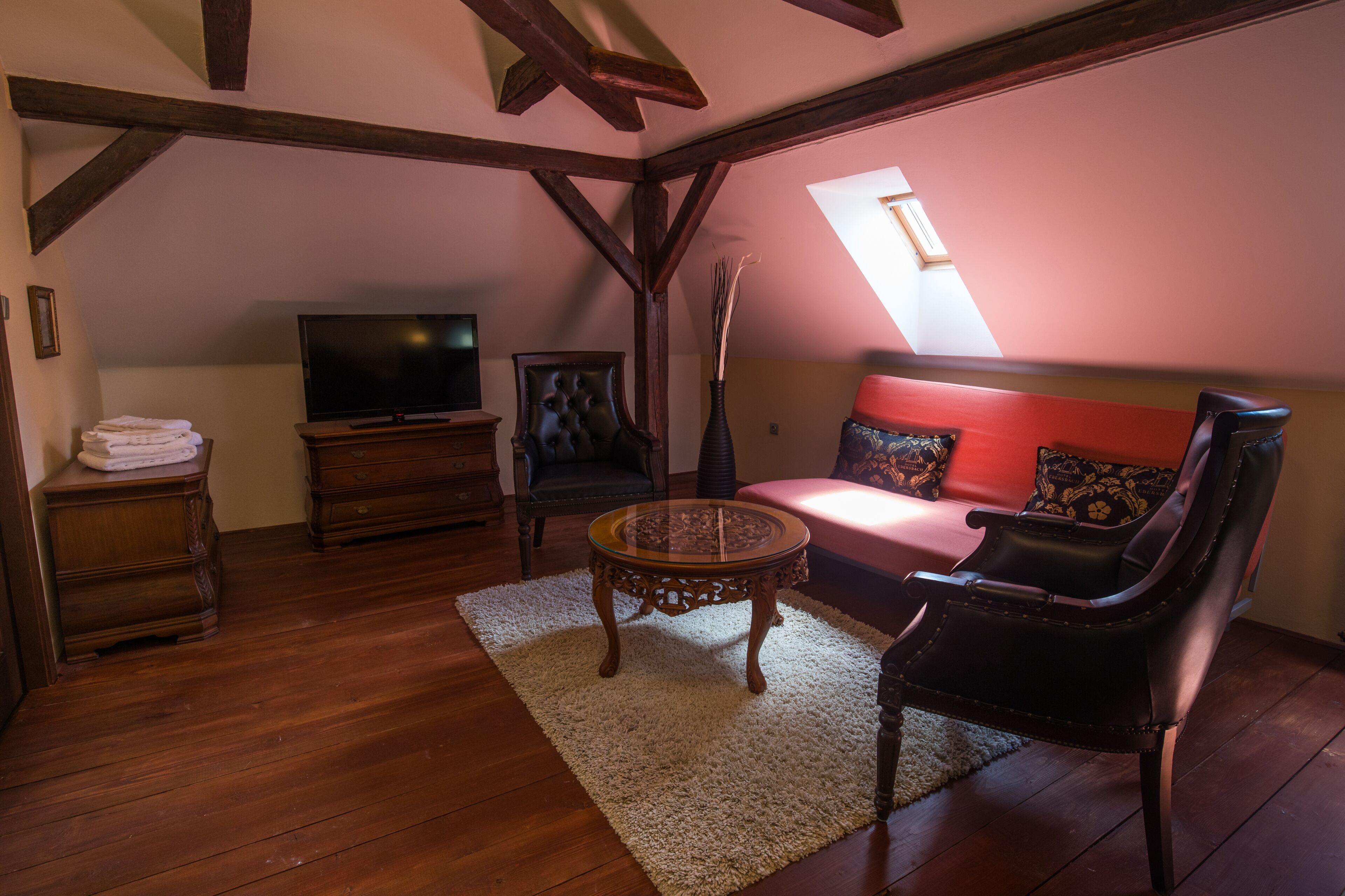 Ein gemütliches Loft-Zimmer mit Holzboden, einem Fernseher, einem Sofa mit roten Kissen, einem schwarzen Ledersessel und einem kleinen runden Tisch.
