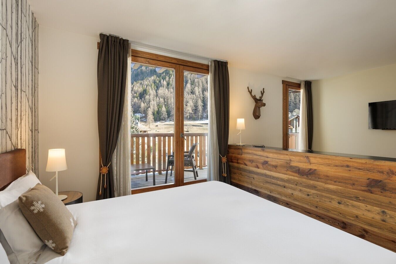 A hotel room with a large bed, a wooden headboard, a balcony with a view of trees and mountains, and a mounted deer head on the wall.