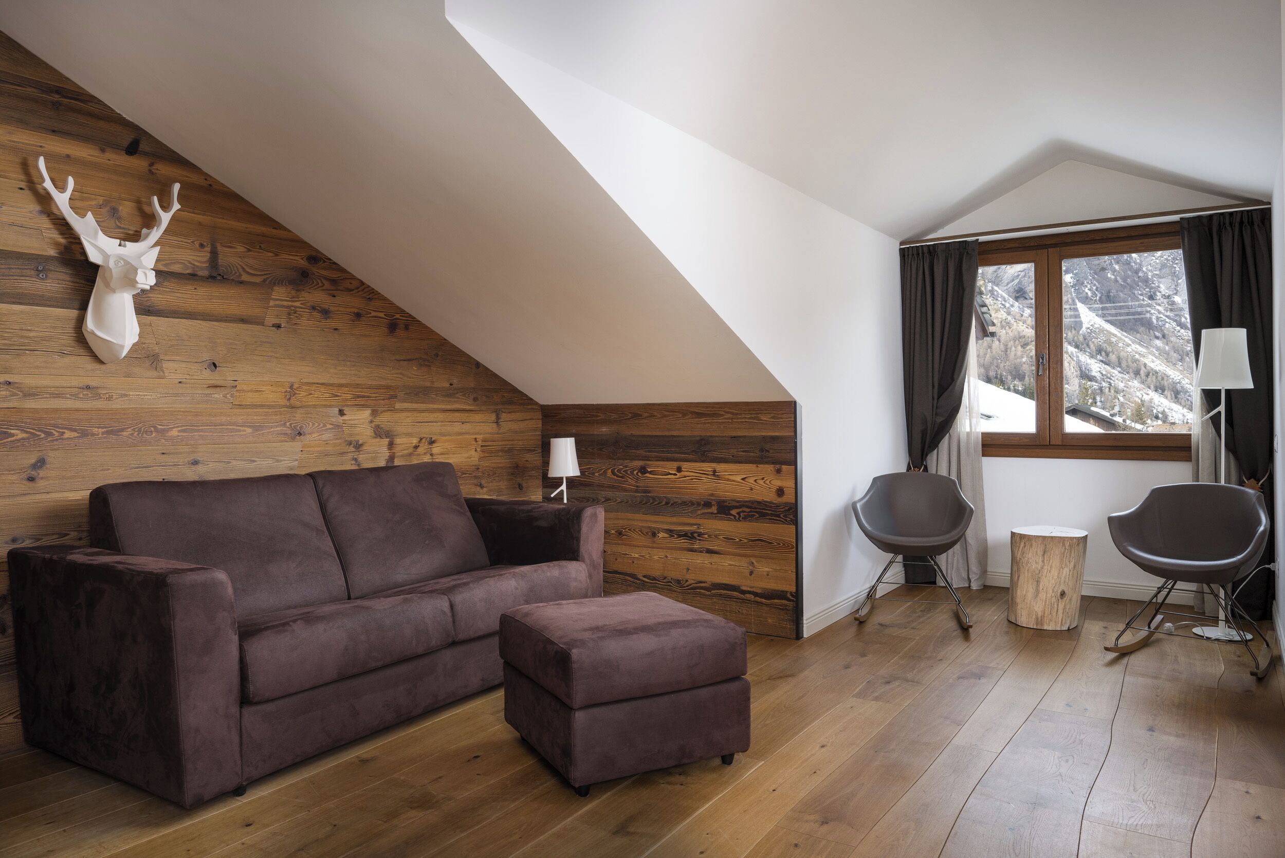 Un salon moderne avec un mur en bois, un canapé marron, un pouf assorti, une lampe, deux fauteuils et une fenêtre avec des rideaux.