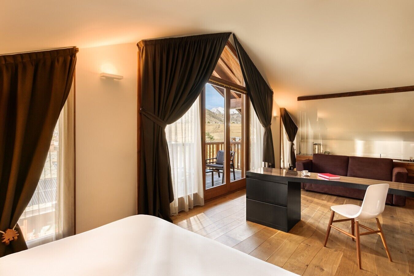 A hotel room with a bed, a desk, a chair, a sofa, and a balcony with a view of the mountains.