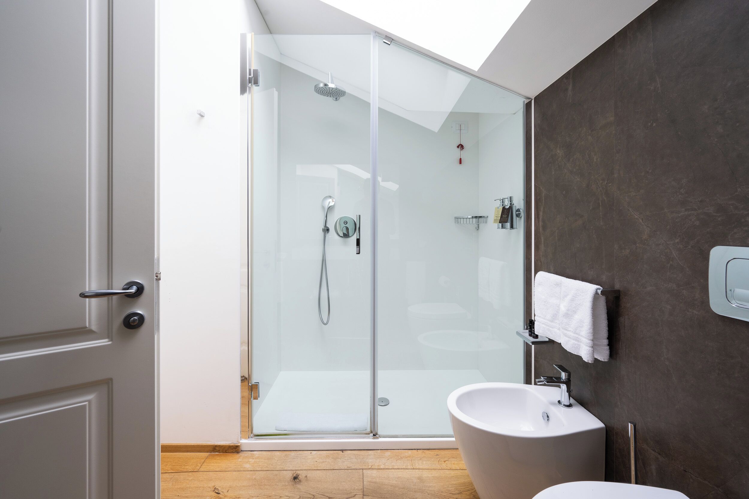 Un baño moderno con cabina de ducha de vidrio, un lavabo blanco y pared con azulejos oscuros.