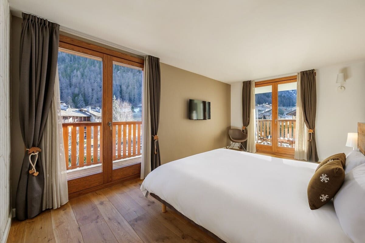 A hotel room with a large bed, wooden flooring, a TV, and a balcony with a view of a snowy landscape.