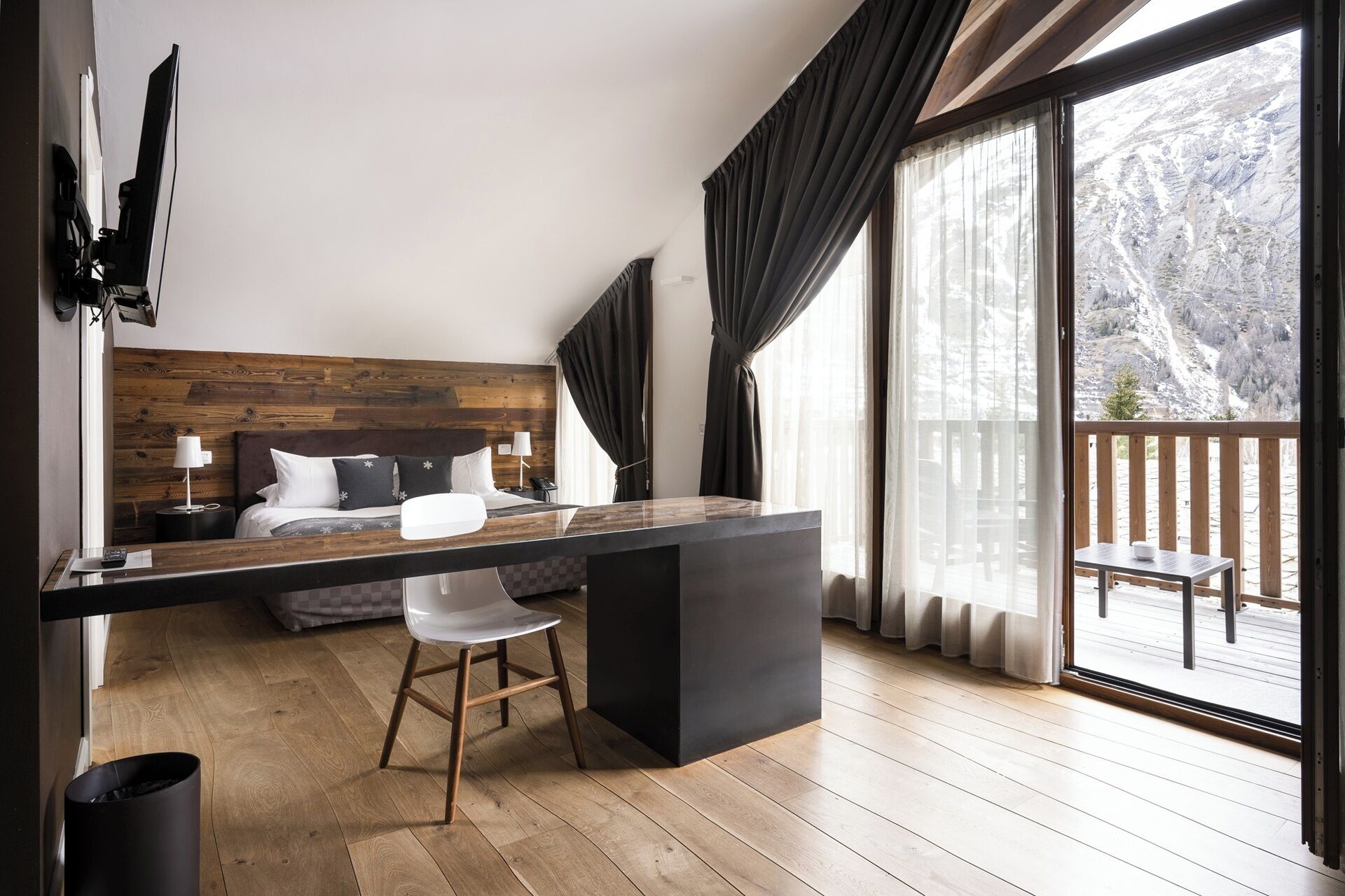 A modern hotel room with a wooden headboard, a large window with curtains, a desk with a chair, and a view of a snowy landscape.
