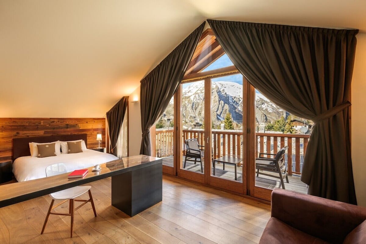 A bedroom with a bed, a desk, and a chair. There is a balcony with a view of mountains.