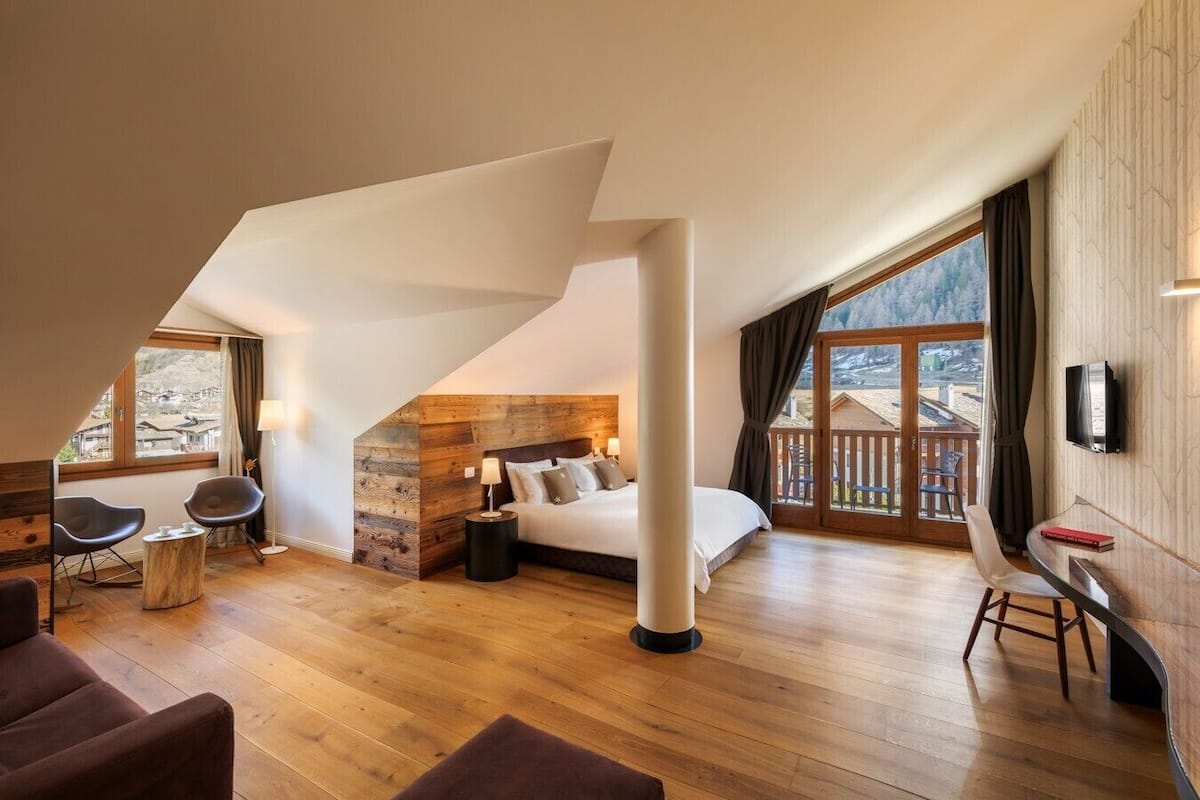 A modern bedroom with a sloped ceiling, wooden walls, a large bed, and a view of a wooden deck outside.