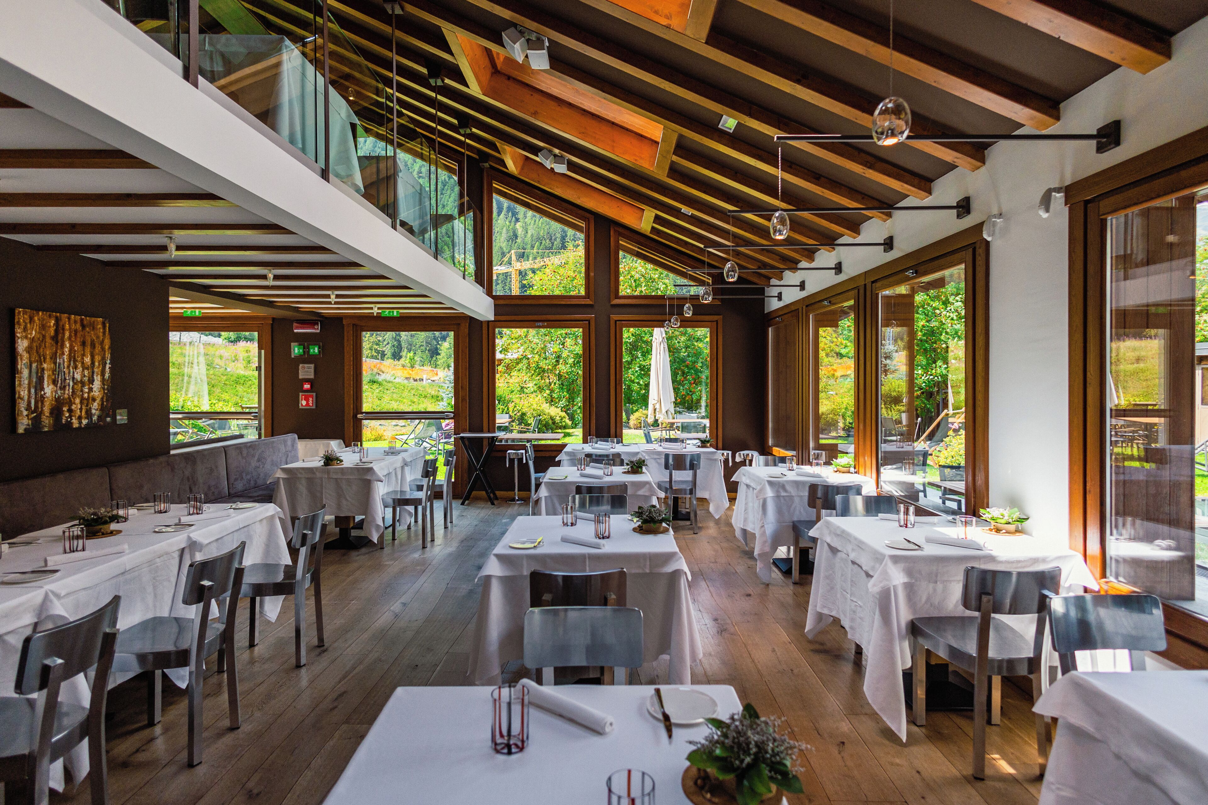 A restaurant with multiple tables, high ceilings, and large windows offering a view of greenery.