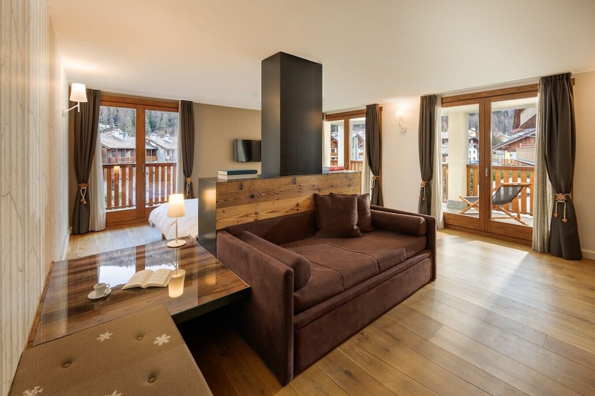 A modern living room with a brown sofa, a wooden coffee table, and a TV on a wooden wall. There are large windows with curtains, a balcony with a wooden railing, and a fireplace.