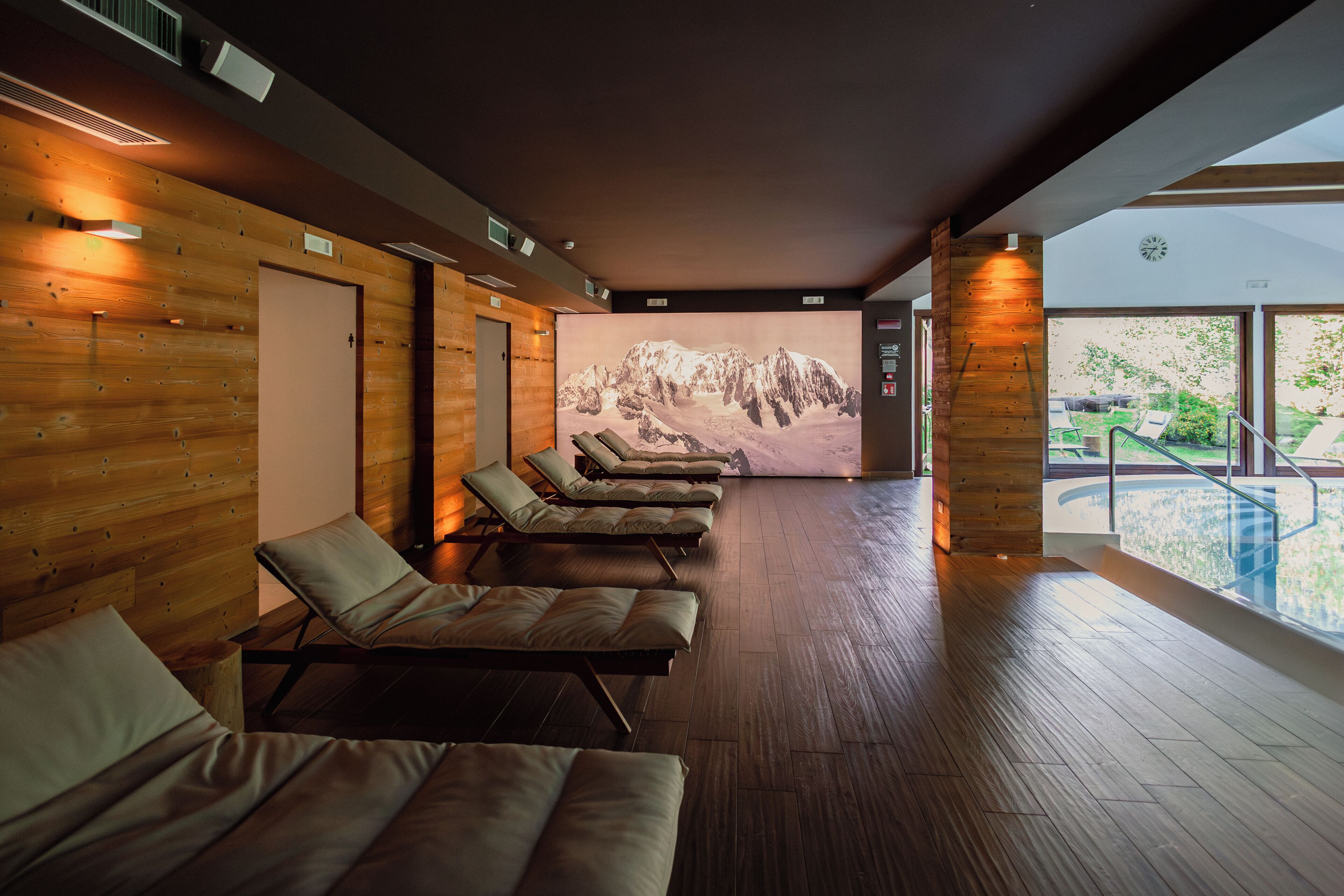 Wellnessbereich mit Holzwänden, einer großen Bergmalerei und einem durch eine Glasscheibe sichtbaren Pool.