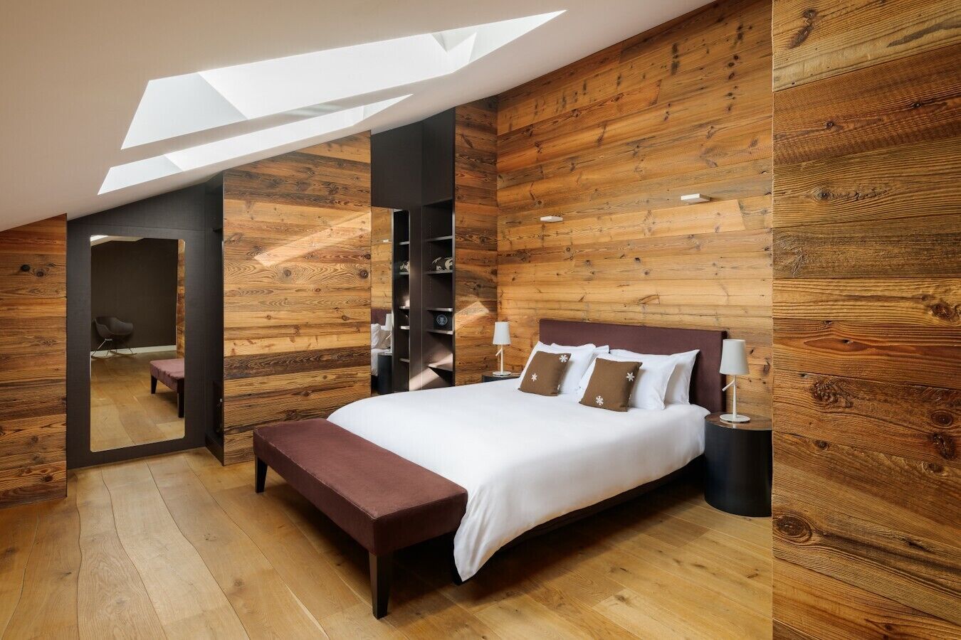 A modern bedroom with a skylight, wooden walls, a large bed with pillows, a bench, and a nightstand with a lamp.